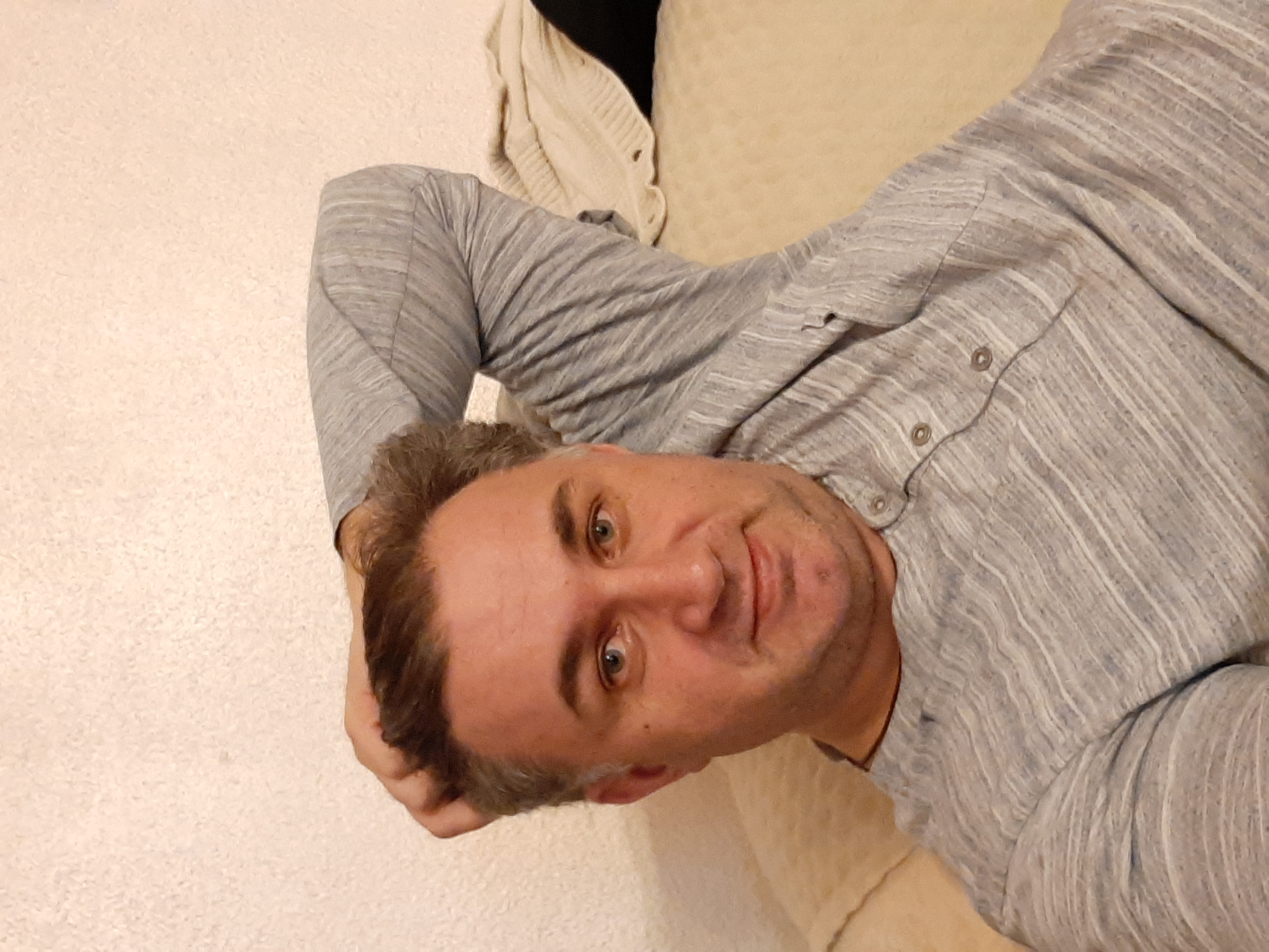 Portret mężczyzny w szarej koszuli leżącego na beżowej sofie, z widocznymi włosami i fragmentem kremowego swetra w tle.