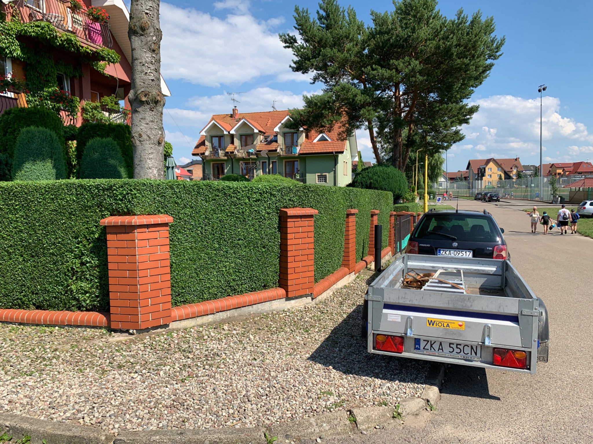 Przycięty żywopłot z ceglanymi słupkami, widoczny z perspektywy ulicy z zaparkowaną przyczepą, domy w tle.