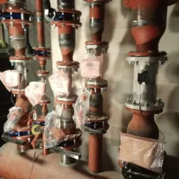 Czerwone rury instalacji grzewczej z zaworami i czujnikami, niektóre elementy zabezpieczone folią bąbelkową, widoczne ślady spawania, tło stanowi ściana z cegieł i palety z materiałami.