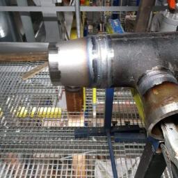 Usługi Hydrauliczno-spawalnicze - Spawany trójnik rur stalowych na ruszcie, widoczne ślady spawania, kolorowe przebarwienia termiczne na spoinie, przemysłowe tło z instalacjami.