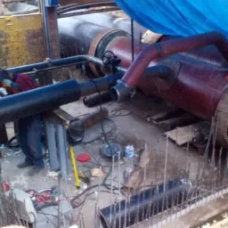 Pracownik w kasku i odblaskowej kamizelce spawa rurę w wykopie, widoczne elementy instalacji, narzędzia, wzmocnienia zbrojeniowe i niebieska plandeka.