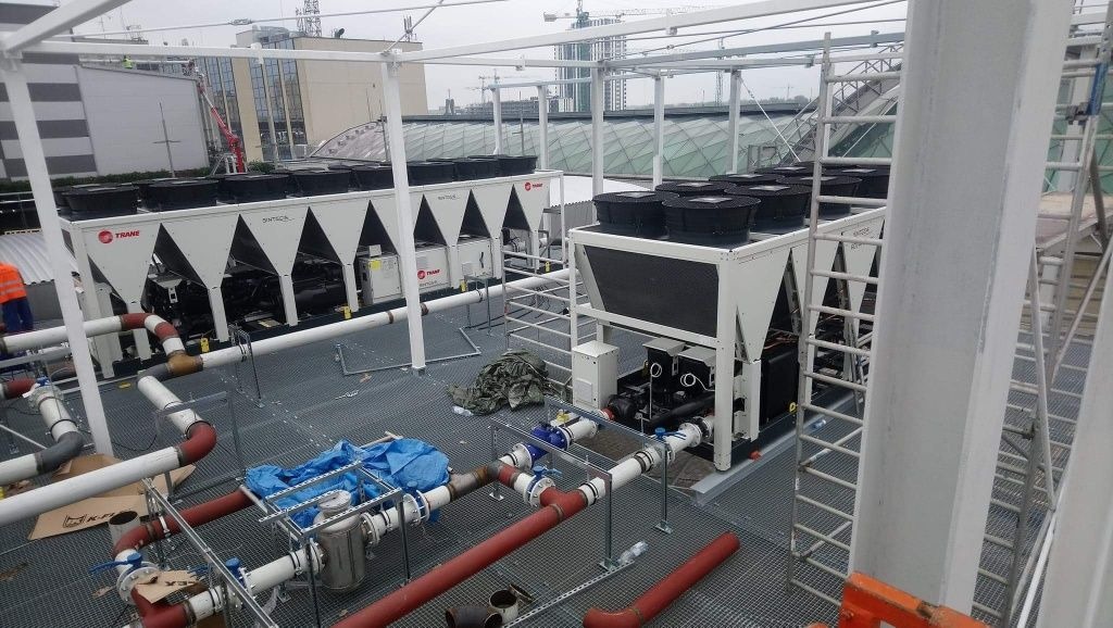 Instalacja przemysłowa na dachu budynku z widocznymi agregatami chłodniczymi Trane, rurociągami w kolorze czerwonym i białym, oraz stalową konstrukcją wsporczą i schodami.
