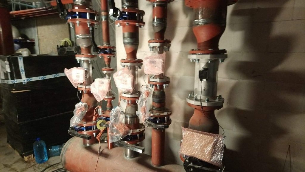 Czerwone rury instalacji grzewczej z zaworami i czujnikami, niektóre elementy zabezpieczone folią bąbelkową, widoczne ślady spawania, tło stanowi ściana z cegieł i palety z materiałami.