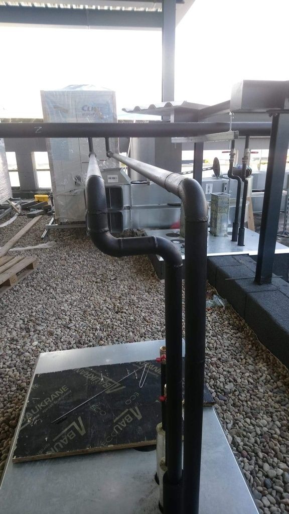 Czarne rury instalacyjne, z widocznymi spawami i izolacją, prowadzone na zewnątrz budynku, z podłożem wysypanym drobnymi kamieniami.