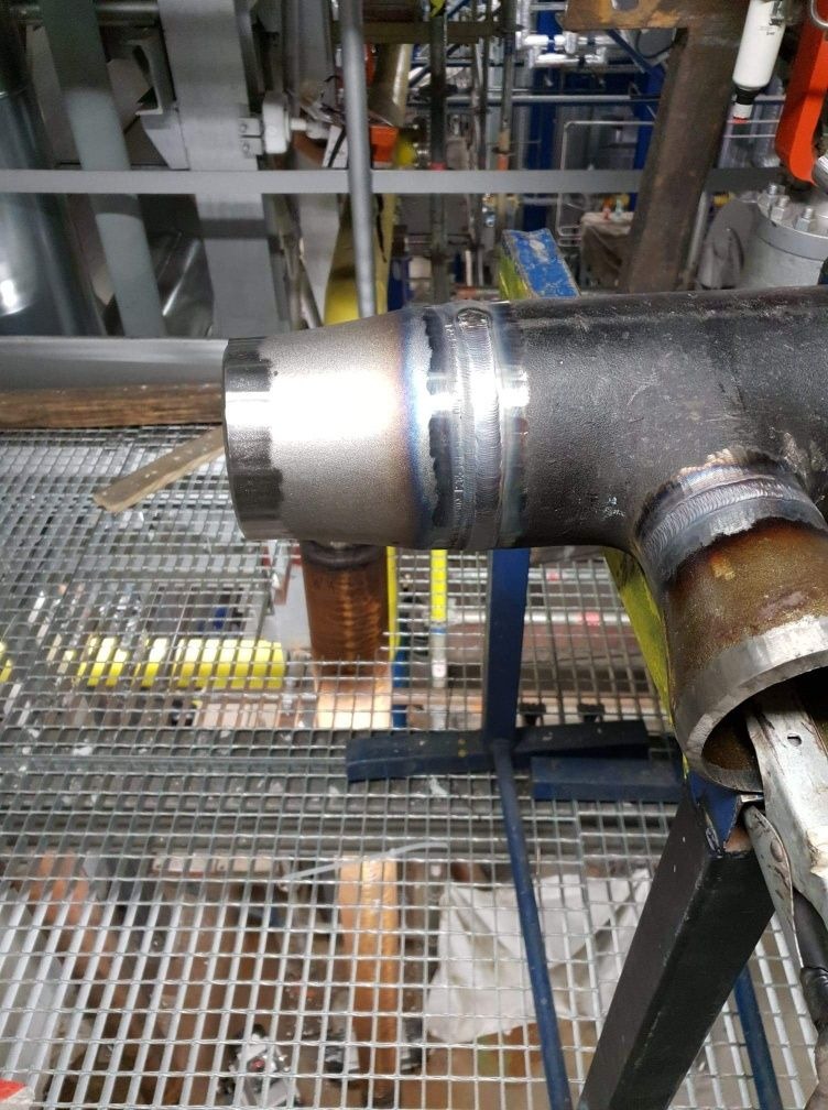 Spawany trójnik rur stalowych na ruszcie, widoczne ślady spawania, kolorowe przebarwienia termiczne na spoinie, przemysłowe tło z instalacjami.