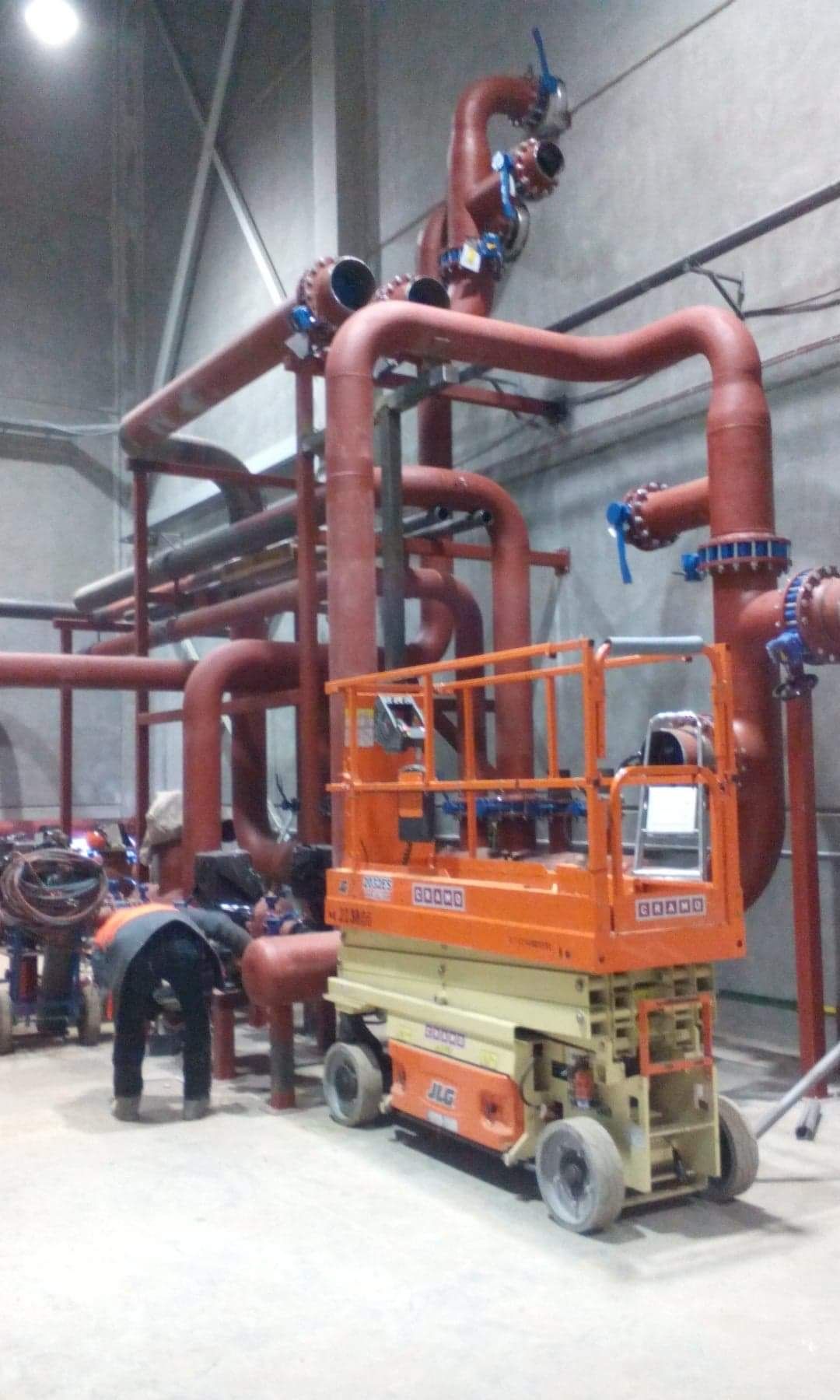 Rozbudowana instalacja rurociągów przemysłowych, malowanych na czerwono, z widocznym podnośnikiem nożycowym JLG i pracownikiem w kamizelce odblaskowej w surowym wnętrzu hali.