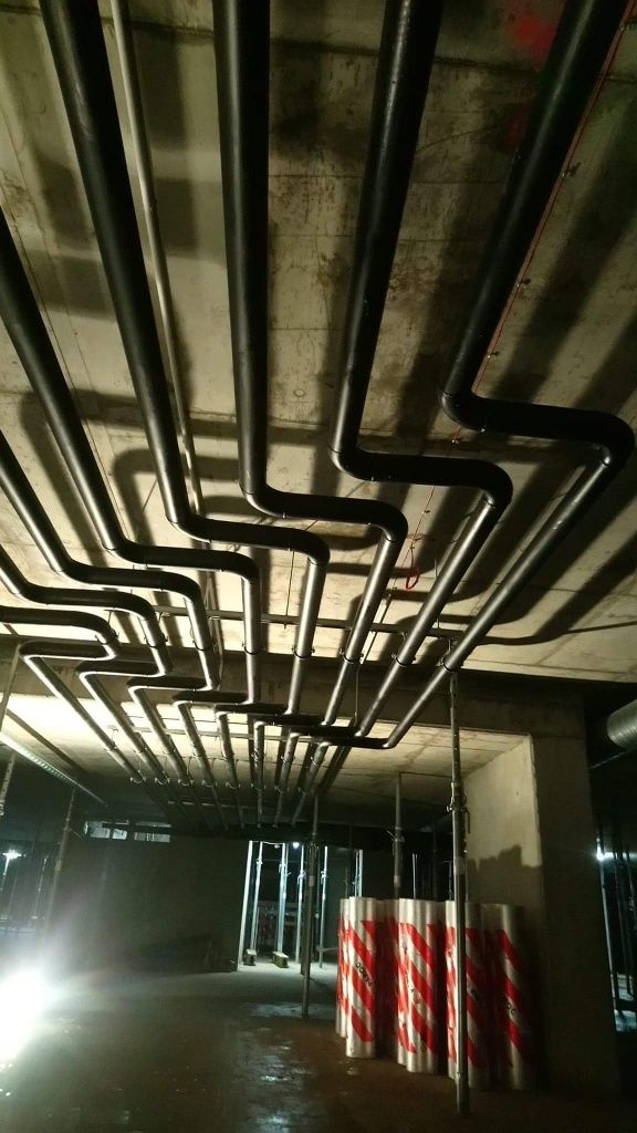 Skomplikowany układ czarnych rur pod betonowym sufitem w surowym wnętrzu, zabezpieczone podpory budowlane z czerwono-białymi pasami w tle, oświetlenie robocze.