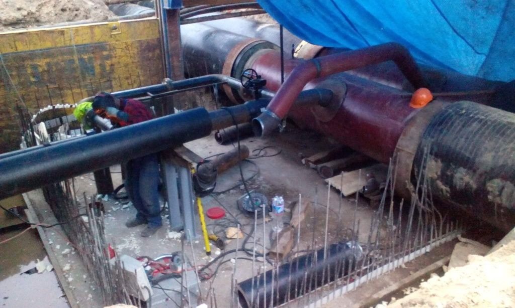 Pracownik w kasku i odblaskowej kamizelce spawa rurę w wykopie, widoczne elementy instalacji, narzędzia, wzmocnienia zbrojeniowe i niebieska plandeka.