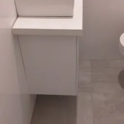 Nowoczesna, kwadratowa umywalka nablatowa z chromowaną baterią, osadzona na białej szafce w minimalistycznej łazience z szarymi płytkami podłogowymi i białymi płytkami ściennymi.