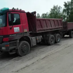 Czerwona ciężarówka z przyczepą na piaszczystym podłożu, widoczne ślady opon, drzewa w tle.