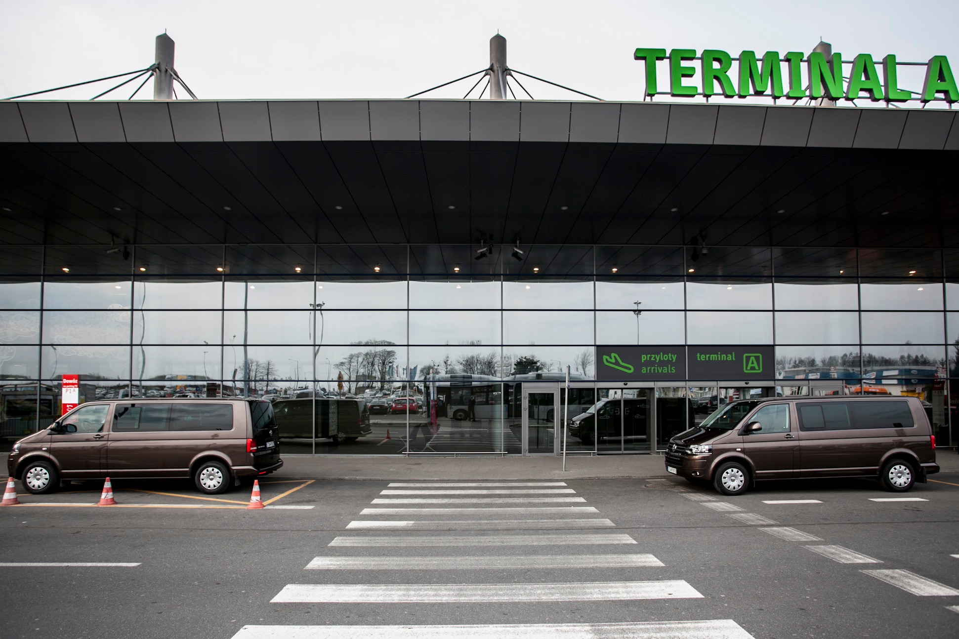 Dwa brązowe busy typu van zaparkowane przed nowoczesnym budynkiem lotniska z napisem 'TERMINAL A' w kolorze zielonym, widoczne przejście dla pieszych i pomarańczowe pachołki.