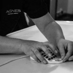 Kobieta w koszulce z logo 'Agnes Fashion Group' używa przyrządu do znakowania wzoru na białym materiale, widoczne dłonie z pierścionkami.