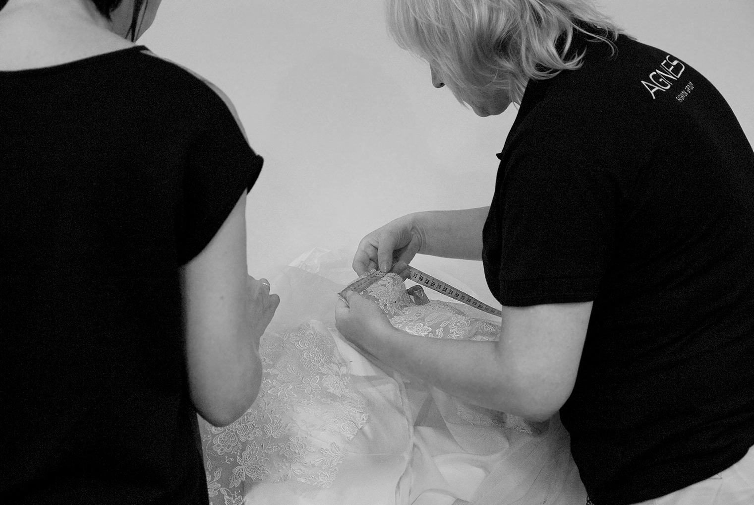 Krawcowa mierzy koronkowy materiał sukni ślubnej za pomocą miarki, widoczny fragment logo 'AGNES' na koszulce pracownika.