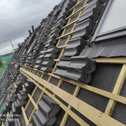 Montaż czarnej dachówki karpiówki na drewnianej konstrukcji z widocznym oknem dachowym i fragmentem rusztowania w tle.