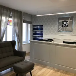 Nowoczesne wnętrze biura z recepcją, sofą i regałem z segregatorami, logo firmy PRO TAX na ścianie, stonowana kolorystyka.