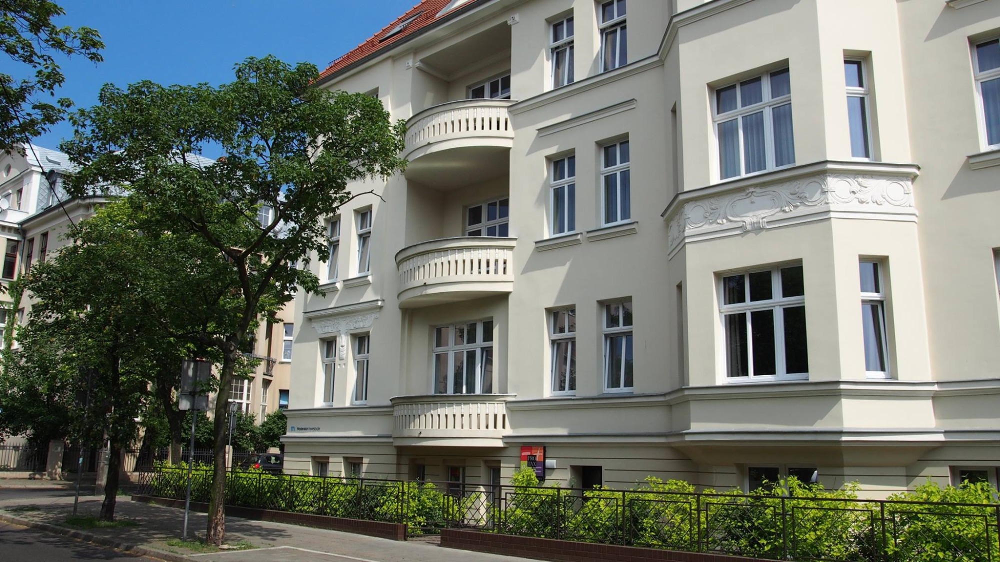 Elewacja odnowionego, kremowego budynku mieszkalnego z balkonami i ozdobnymi detalami architektonicznymi, widok z poziomu ulicy, z zielenią i drzewem w pierwszym planie.