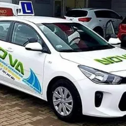 Biały samochód z zielonym napisem NOVA i niebieską grafiką fali, oznaczony literą 'L' na dachu, zaparkowany przed budynkiem.