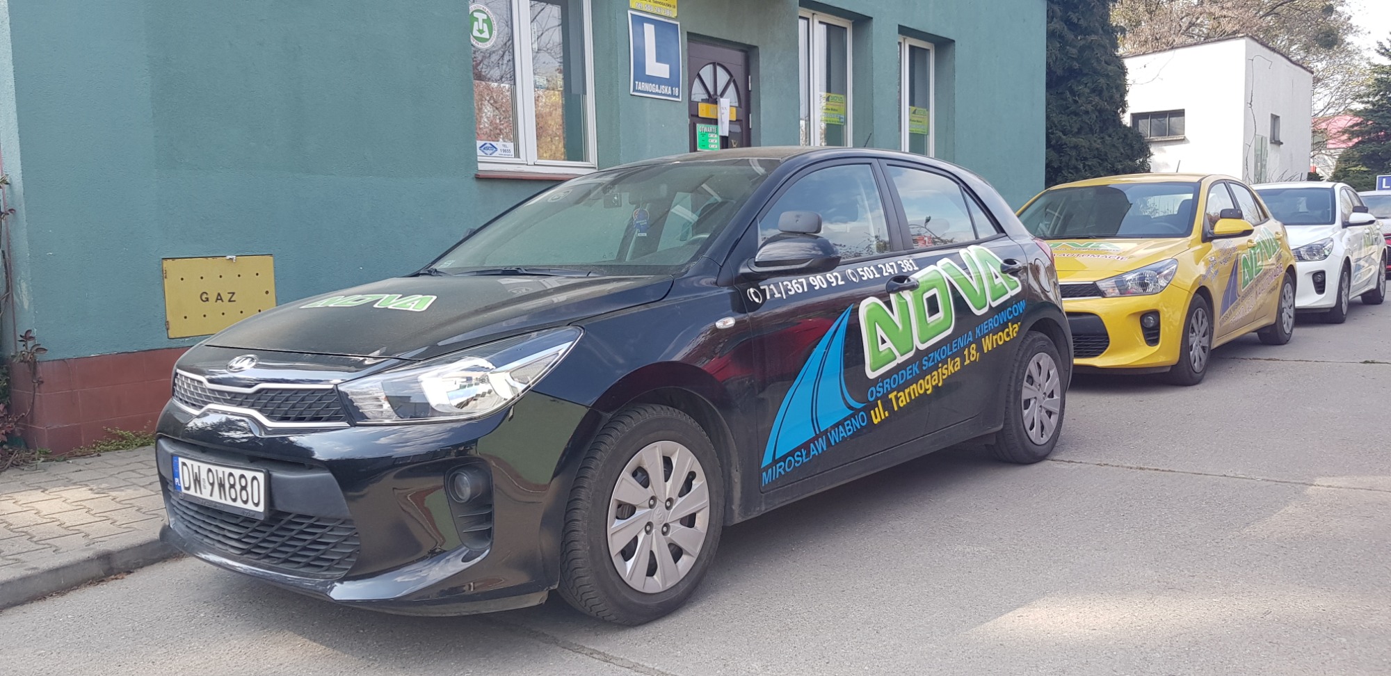 Szereg samochodów z logo i numerami telefonów ośrodka szkolenia kierowców zaparkowanych przed budynkiem z szyldem 'L' i adresem Tarnogajska 18.