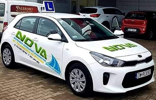 Biały samochód z zielonym napisem NOVA i niebieską grafiką fali, oznaczony literą 'L' na dachu, zaparkowany przed budynkiem.