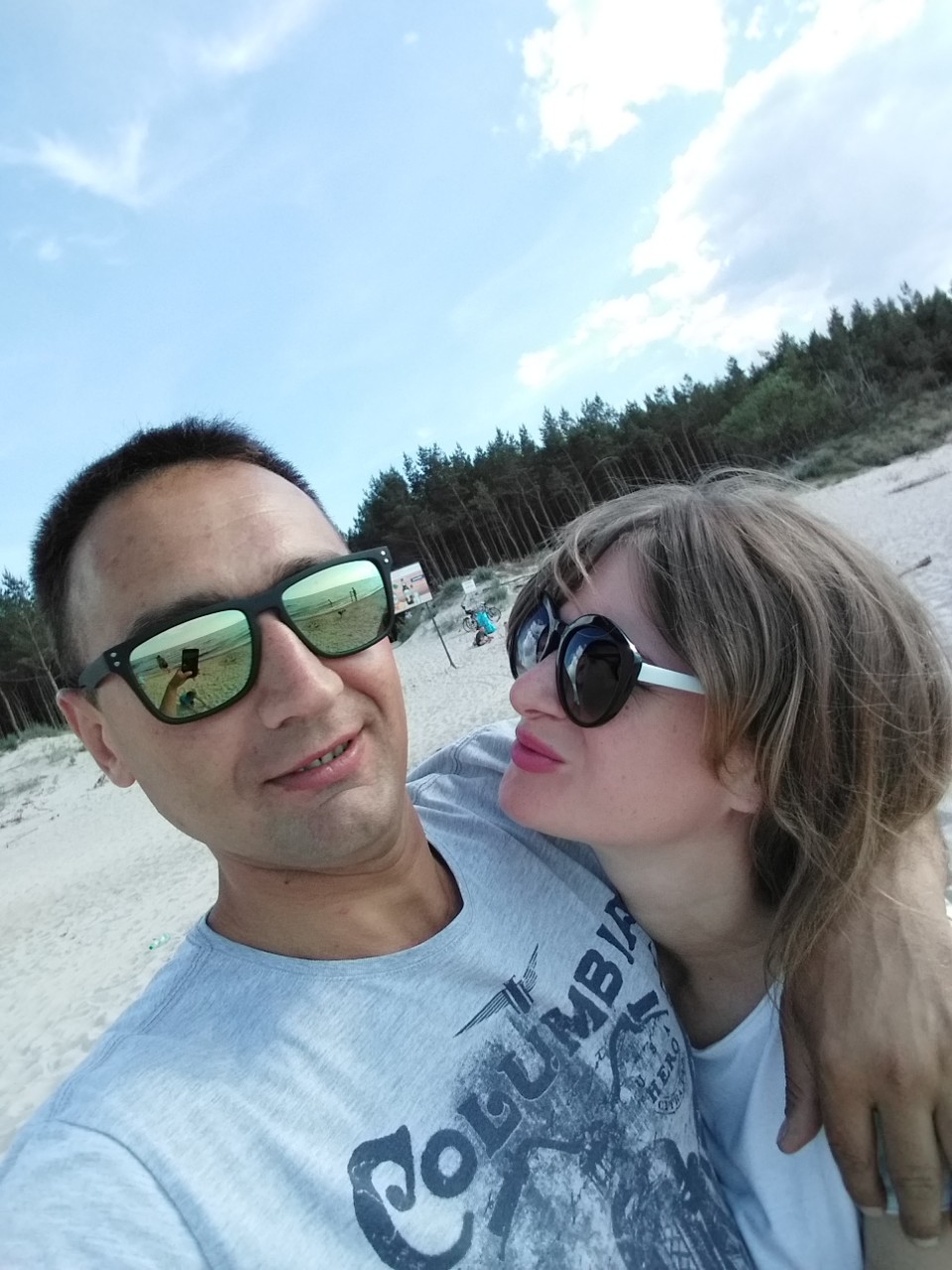 Para na plaży w okularach przeciwsłonecznych, w odbiciu w okularach mężczyzny widać plażę i osoby.