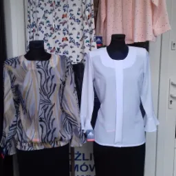 Wystawa sklepowa z damską odzieżą: bluzki z długim rękawem na manekinach i wieszakach, wzory w motyle, abstrakcyjne fale i gładka różowa, zestawione z czarnymi spódnicami.