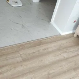 Połączenie podłogi z paneli w odcieniu jasnego drewna z marmurową podłogą, widoczna listwa progowa, w tle biała szafka nocna z rośliną.