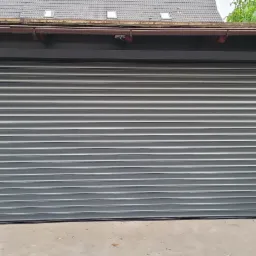 Zewnętrzna, szara, metalowa roleta antywłamaniowa, zamontowana na garażu, widoczny fragment dachu i numer posesji na sąsiednim ogrodzeniu.