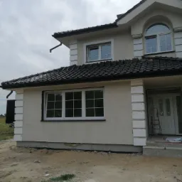 Dwukondygnacyjny dom z jasną elewacją, białymi zdobieniami narożników i ciemną dachówką, widoczny fragment terenu przed budynkiem w trakcie prac wykończeniowych.