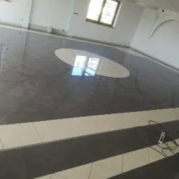 Wykończona posadzka z ciemnoszarych i kremowych płytek w dużym, jasnym pomieszczeniu z białymi ścianami i łukowymi elementami dekoracyjnymi; widoczne okno z odbiciem drzew i przedłużacz na podłodze.