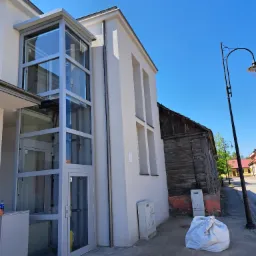 Zewnętrzna przeszklona winda przy budynku z białą elewacją, widok z dołu, obok budynek z drewnianą elewacją, lampa uliczna i worek z odpadami.