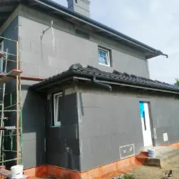 Dom jednorodzinny w trakcie ocieplania elewacji styropianem grafitowym, widoczne rusztowanie, fragment dachu z dachówką ceramiczną i przygotowane podłoże gruntowe wokół budynku.