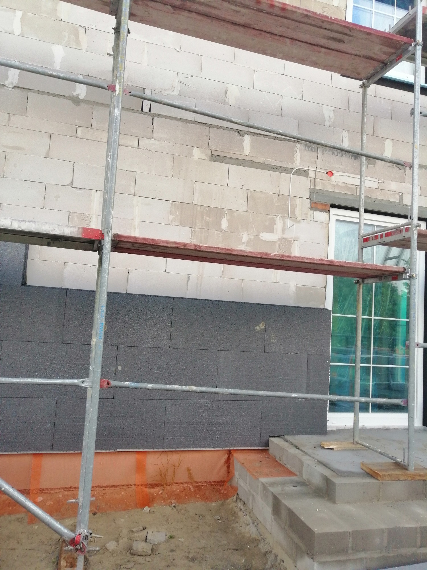 Montaż grafitowego styropianu na elewacji budynku z białego bloczka, widoczne rusztowanie i pomarańczowa membrana w gruncie.