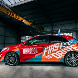 Oznakowany samochód z literą 'L' na dachu, należący do firmy z Krzyża Wielkopolskiego, zaparkowany w hali z suwnicą, z widocznym logo 'AKROPOL pierwszy krok do prawdziwej motoryzacji' i napisem...