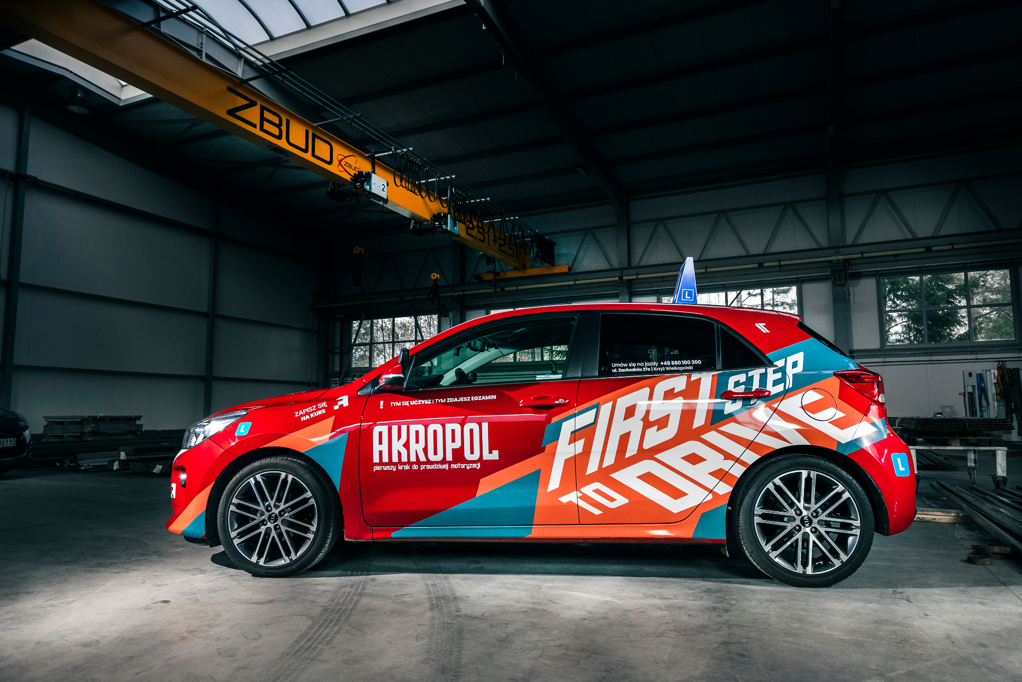 Oznakowany samochód z literą 'L' na dachu, należący do firmy z Krzyża Wielkopolskiego, zaparkowany w hali z suwnicą, z widocznym logo 'AKROPOL pierwszy krok do prawdziwej motoryzacji' i napisem...