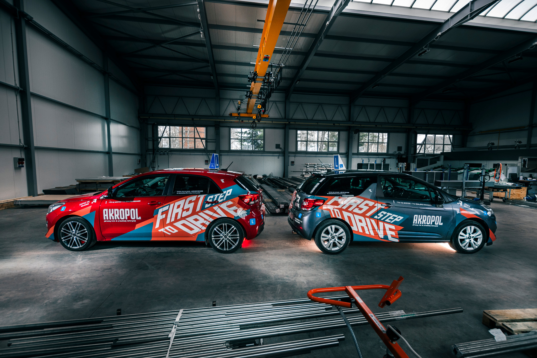 Dwa samochody z oznaczeniami firmy nauki jazdy, czerwony i szary, zaparkowane w hali z suwnicą i plikami rur.