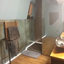 Nowoczesna łazienka z podświetleniem LED wzdłuż podłogi i pionowej ściany prysznica. Na pierwszym planie drewniany grzejnik drabinkowy z szarymi ręcznikami. Ściany wykończone płytkami imitującymi...