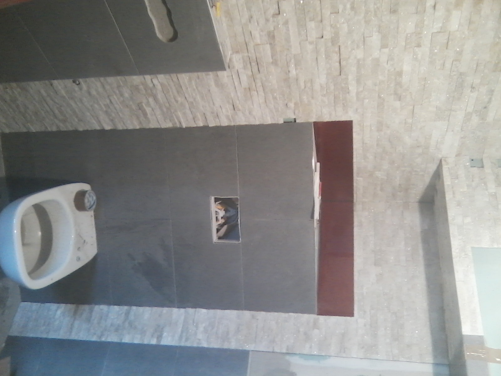 Nowa toaleta podwieszana w trakcie montażu w łazience w Bytomiu, szare płytki na ścianach i podłodze, dekoracyjna ściana z małych, jasnych płytek. Widoczny otwór w ścianie na instalację.