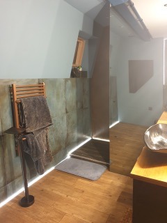 Nowoczesna łazienka z podświetleniem LED wzdłuż podłogi i pionowej ściany prysznica. Na pierwszym planie drewniany grzejnik drabinkowy z szarymi ręcznikami. Ściany wykończone płytkami imitującymi...