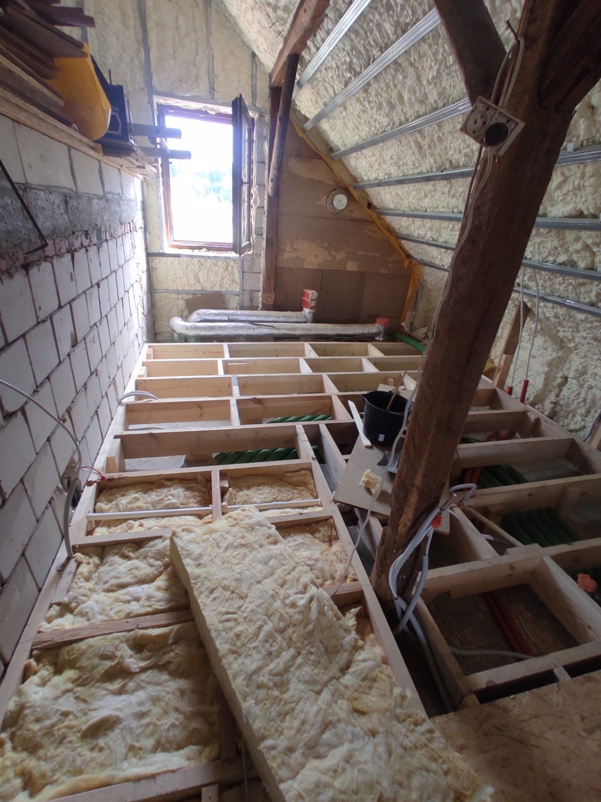 Ocieplenie poddasza wełną mineralną, widoczne krokwie, instalacja elektryczna i grzewcza oraz okno dachowe w trakcie prac wykończeniowych.