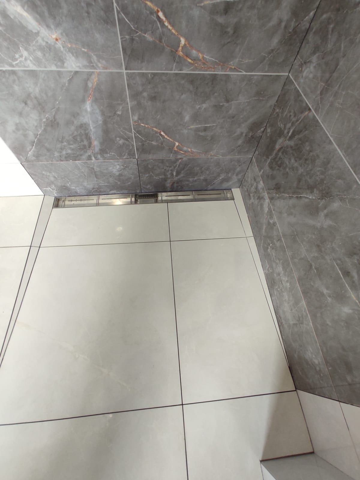 Wykończona łazienka z szarymi płytkami imitującymi marmur na ścianach i jasnymi płytkami na podłodze; widoczny odpływ liniowy.