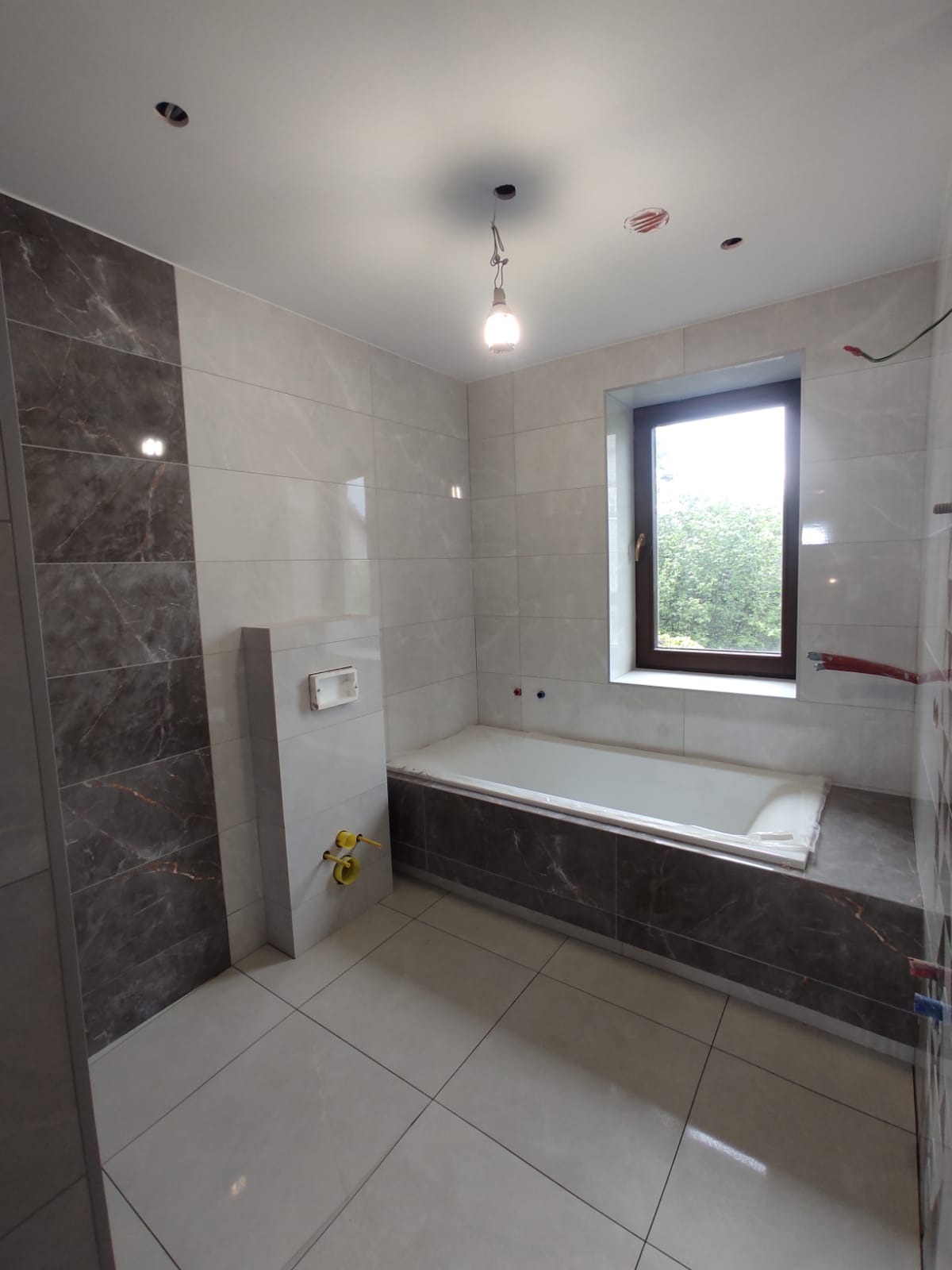 Wnętrze łazienki w trakcie prac wykończeniowych, widoczna obudowana wanna z marmurowym wzorem, jasne płytki na ścianach i podłodze, przygotowane instalacje wodne, surowa żarówka na kablu, okno...