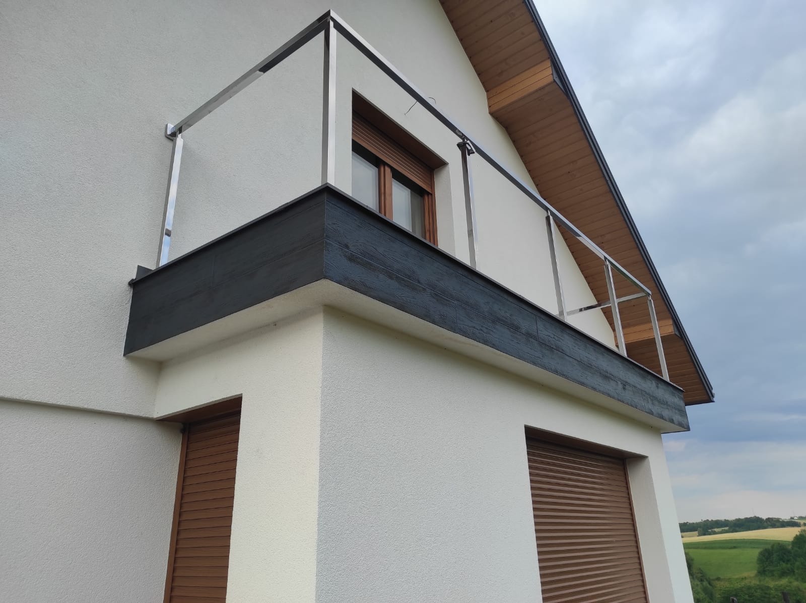 Elewacja domu z balkonem zabezpieczonym metalową balustradą, widoczne okna z brązowymi roletami zewnętrznymi i fragment dachu.