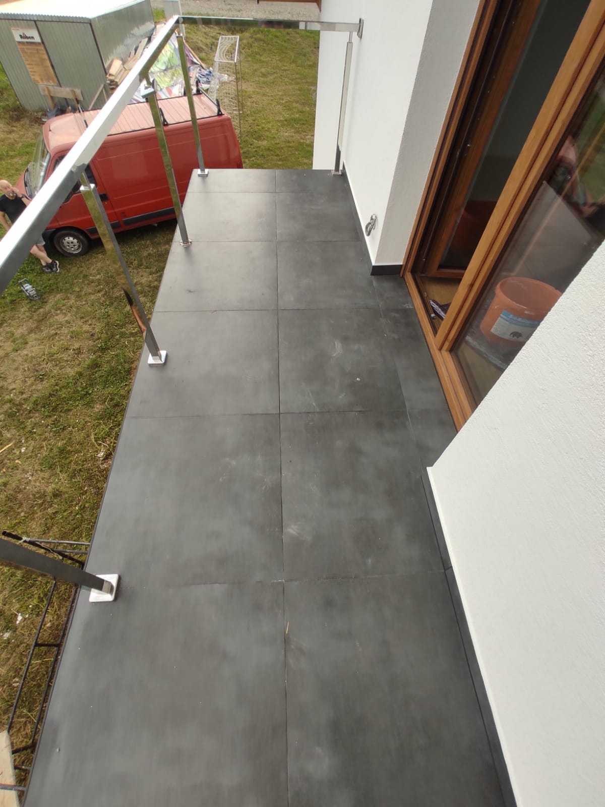 Wykończony balkon z szarymi płytkami, stalową balustradą i widokiem na czerwony samochód dostawczy oraz budynek gospodarczy.