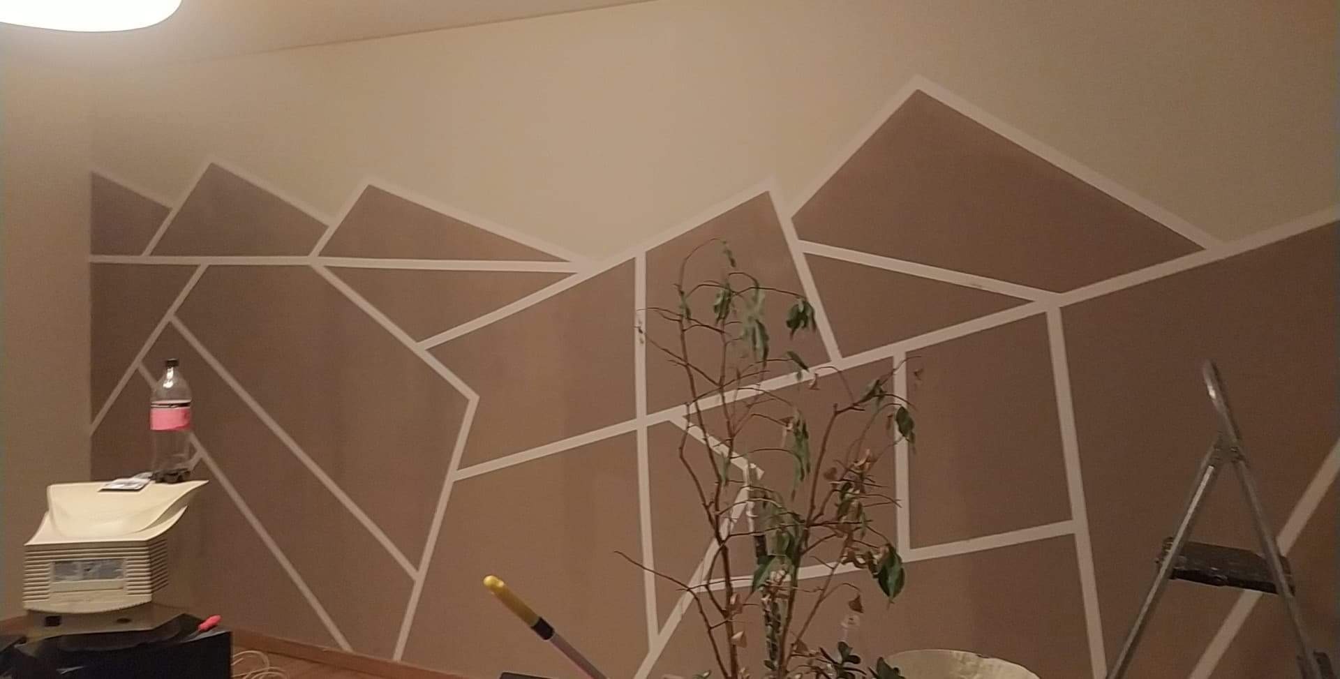 Ściana pomalowana w geometryczne wzory w odcieniach beżu i brązu, obklejona białą taśmą malarską, z widoczną butelką wody, rośliną doniczkową i drabiną.
