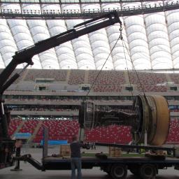 Mar-Trans - Ciężki dźwig samochodowy podnosi turbinę silnika odrzutowego na tle trybun stadionu