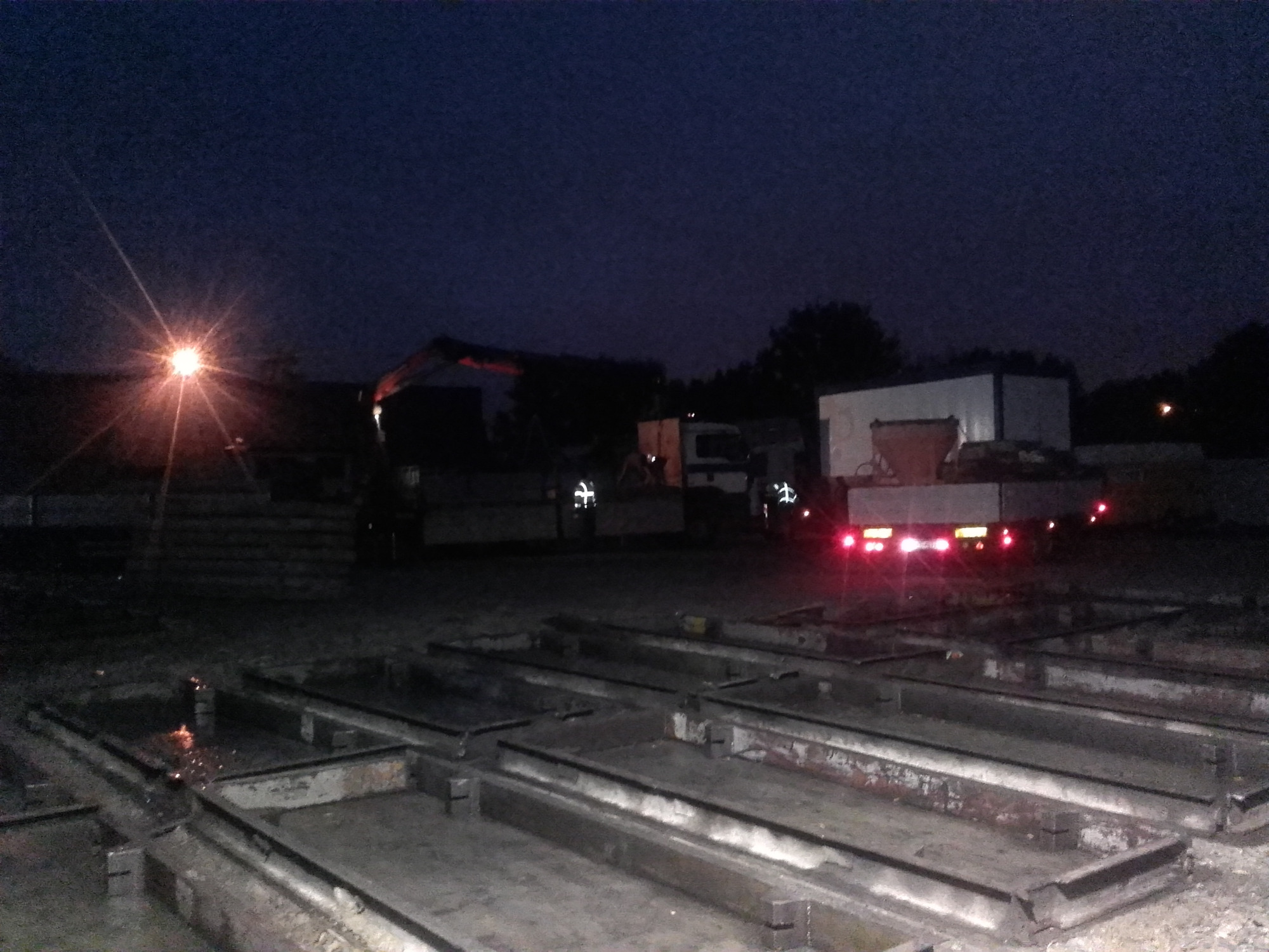 Nocny widok placu budowy z ciężarówkami w tle, oświetlonego pojedynczą lampą, z formami do odlewów betonowych na pierwszym planie.