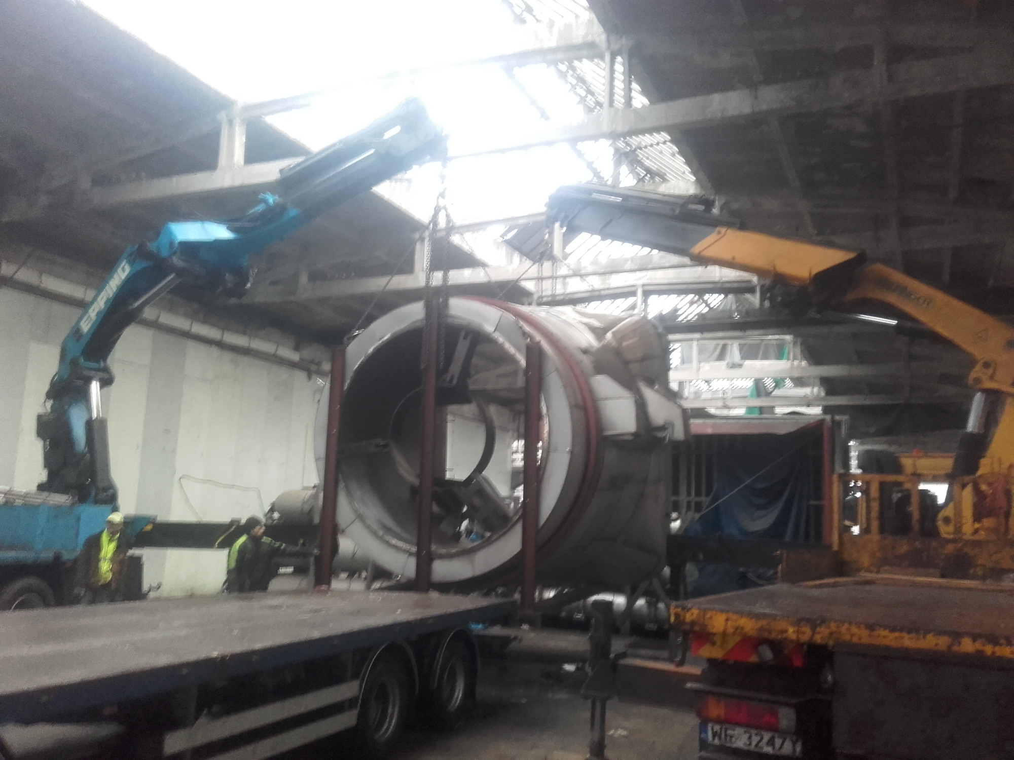 Dwa dźwigi, jeden niebieski EFFER i jeden żółty PEUGEOT, podnoszą duży, okrągły element konstrukcyjny z metalu na naczepę ciężarówki w hali oświetlonej przez dziurawy dach.