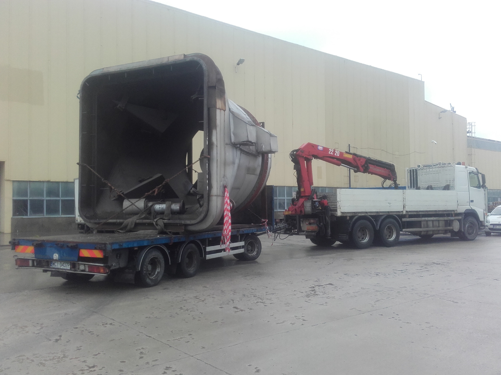 Nietypowy ładunek o dużych gabarytach, przypominający element przemysłowy, zabezpieczony na niskopodwoziowej naczepie ciężarówki z dźwigiem hydraulicznym na tle hali przemysłowej.