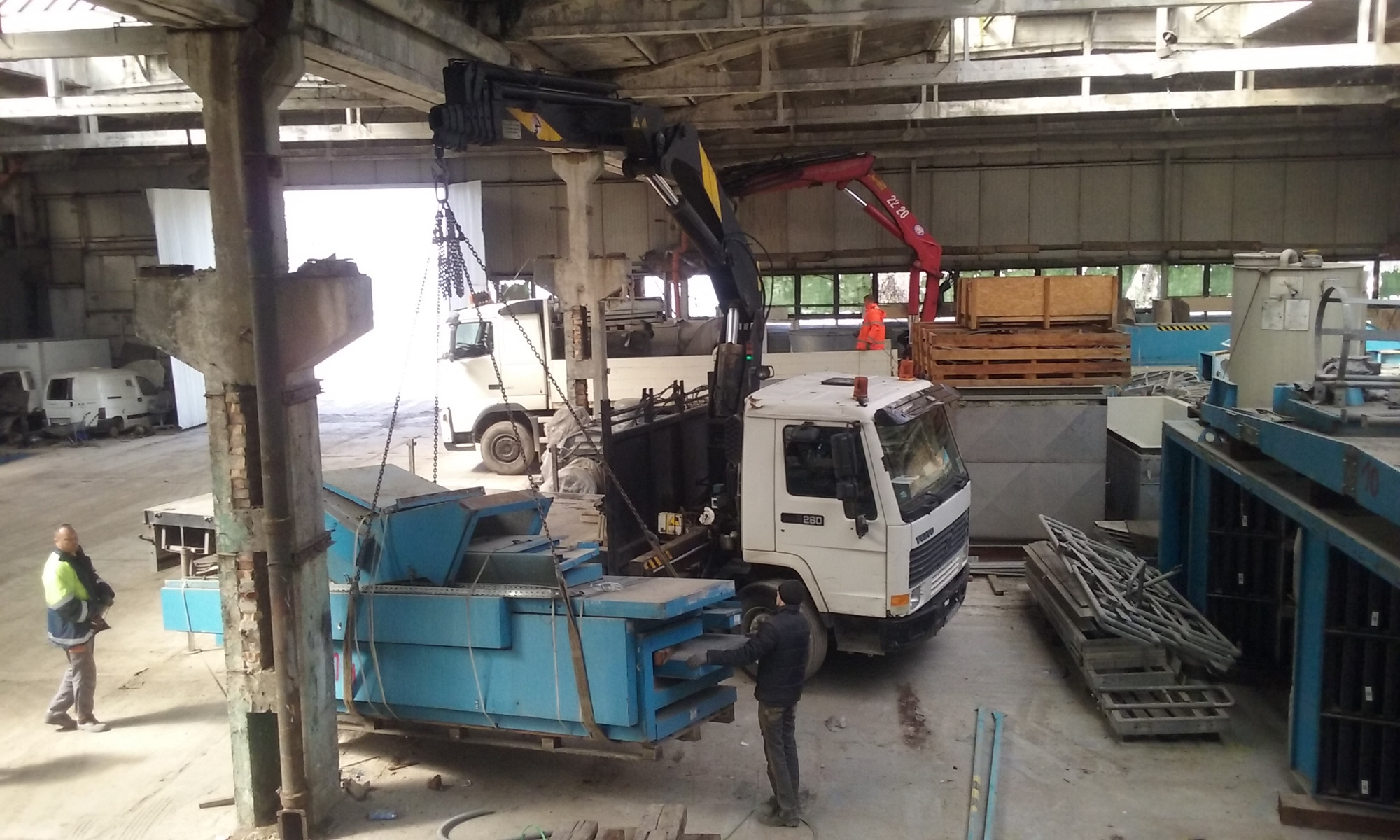 Ciężarówka z dźwigiem HDS załadowuje niebieski, metalowy kontener wewnątrz hali magazynowej; widoczni pracownicy i inne pojazdy.