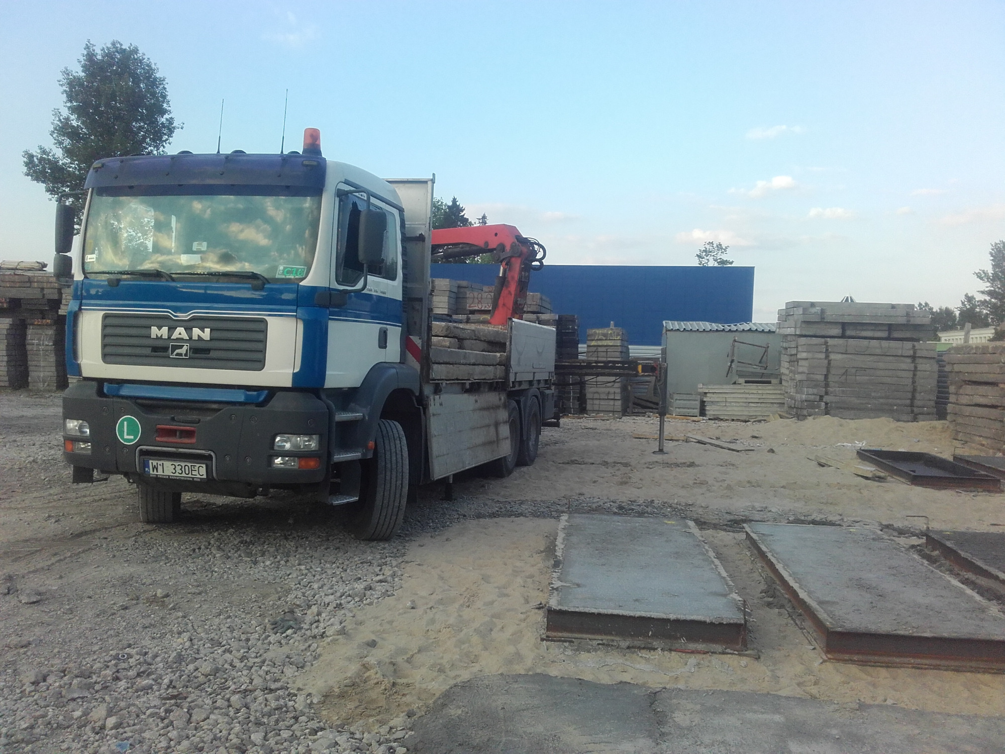 Ciężarówka MAN z czerwoną dźwignią załadowana betonowymi płytami na terenie składu budowlanego, widoczne zapasy płyt i konstrukcji betonowych.
